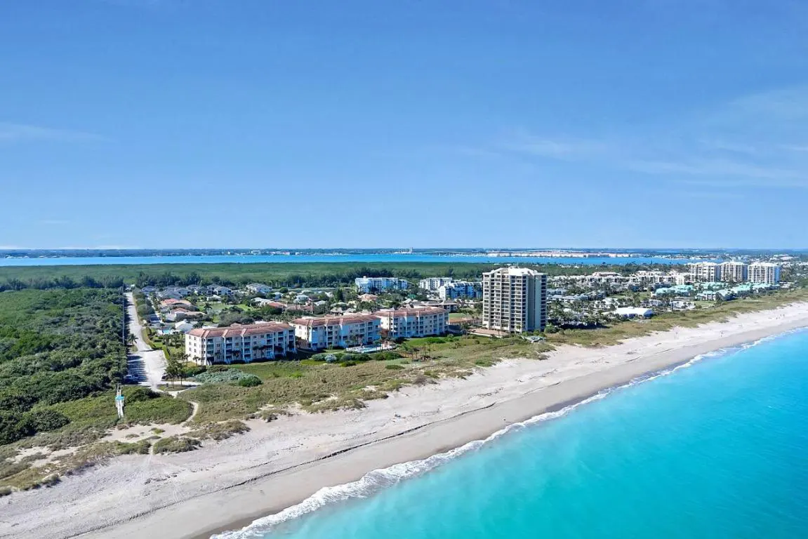 Aerial view of a beach with turquoise water, white sand, and buildings under a clear blue sky.