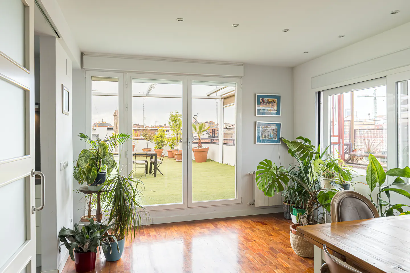 Bright living room with wood floors, plants, and a terrace. The terrace has green turf, potted plants, and a table with chairs.