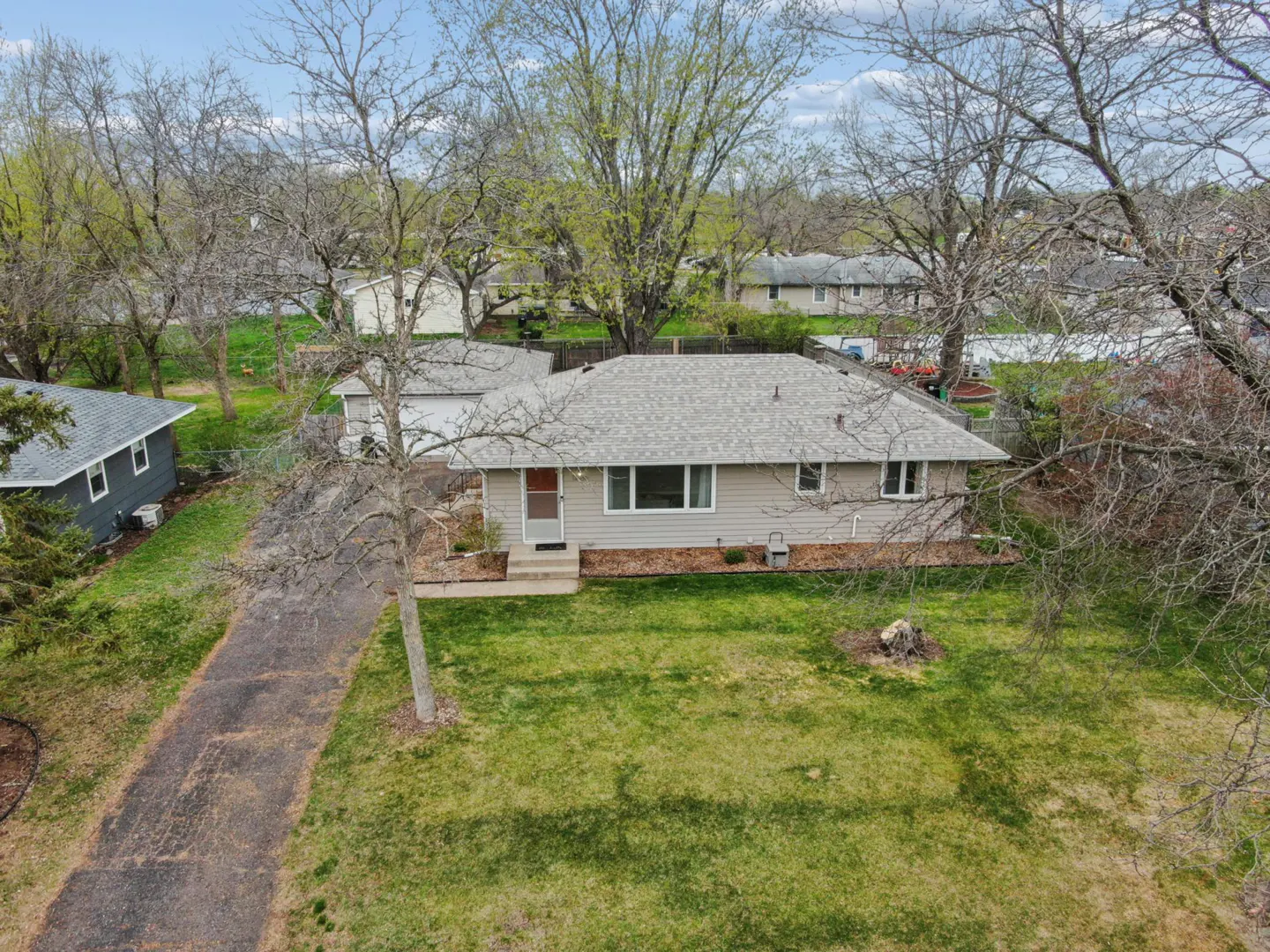 Aerial view of a one-story, light-gray house with a gray roof and a red front door, surrounded by green lawn and trees.