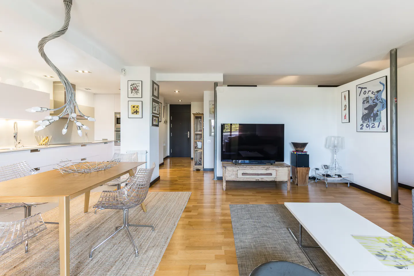 Bright, modern living space with wood floors, a dining table with clear chairs, and a large TV. Artwork adorns the white walls.