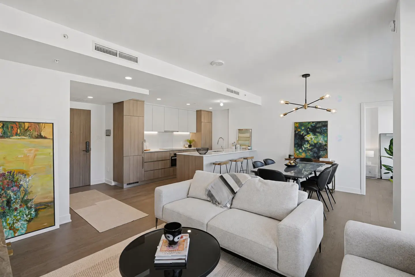 Modern open-concept living area with a white sofa, round coffee table, dining set, and kitchen with light wood cabinets.