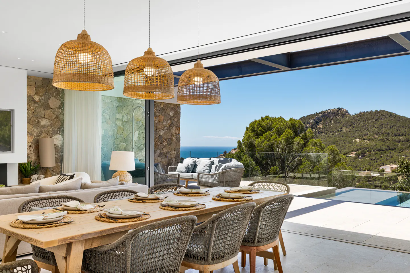 A bright dining room with a long wooden table set for a meal, wicker chairs, and pendant lights. Outside, a view of the ocean and mountains.