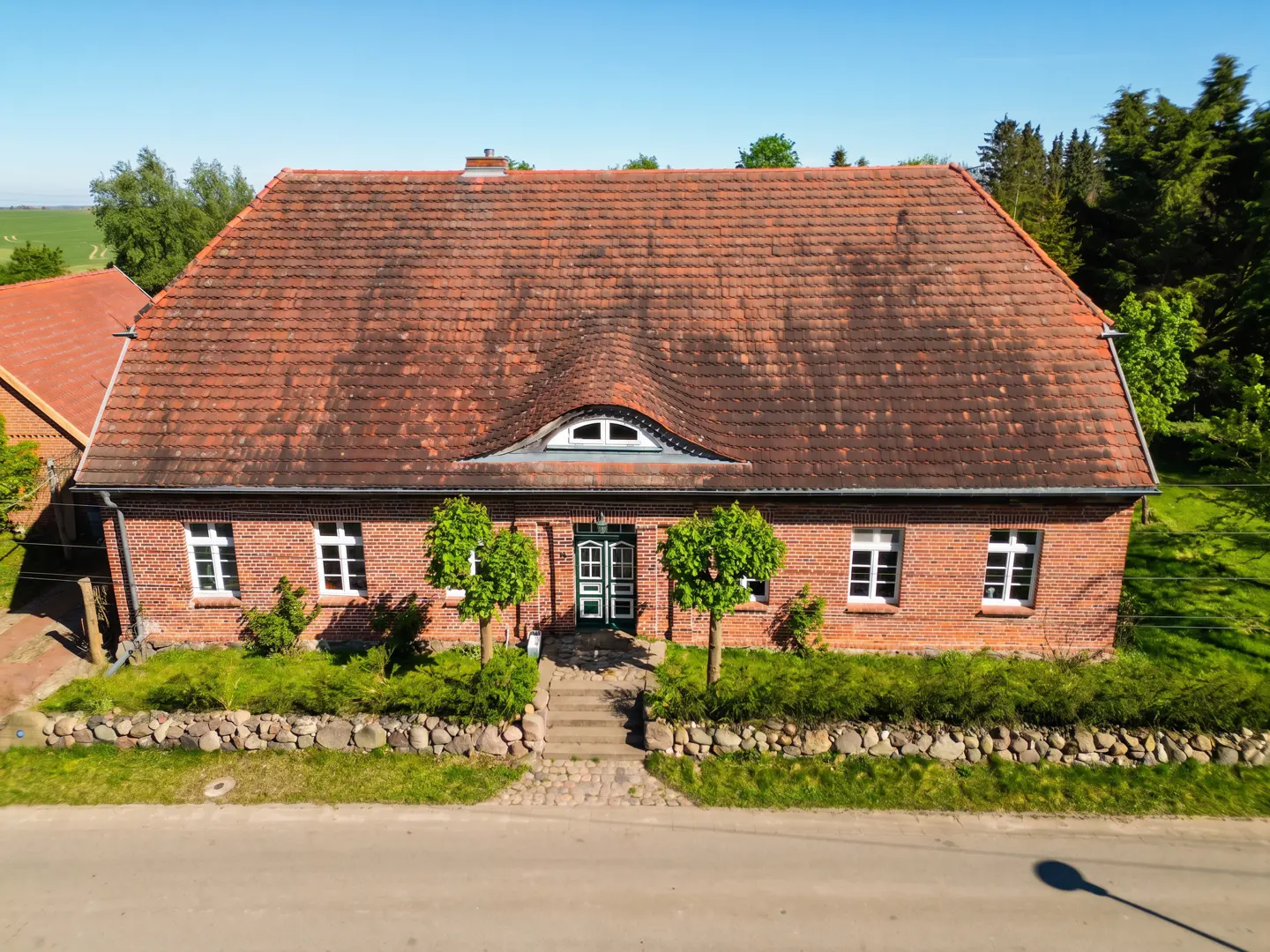 Two-story brick house with a red tile roof, white windows, and a green door. A stone wall and green bushes line the front.