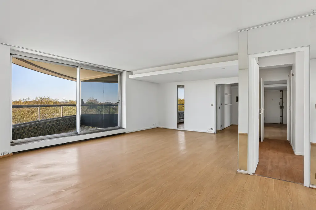 Bright, empty apartment with wood floors and white walls. A large sliding glass door leads to a balcony with a view of trees.