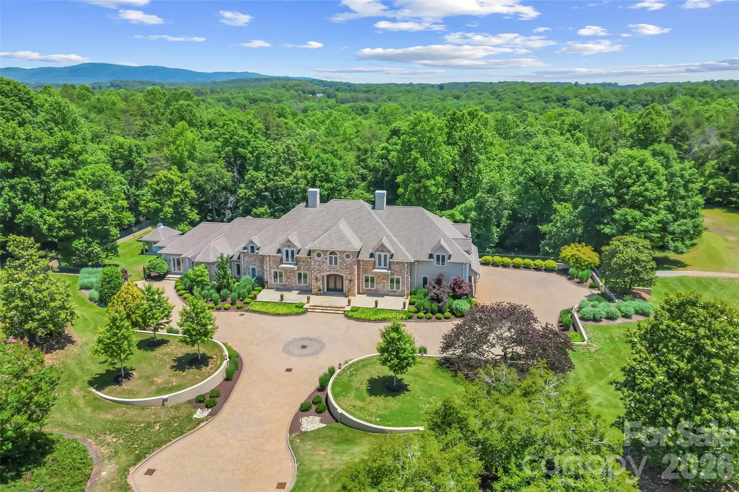Aerial view of a large stone mansion with a circular driveway, surrounded by lush green trees and distant mountains.