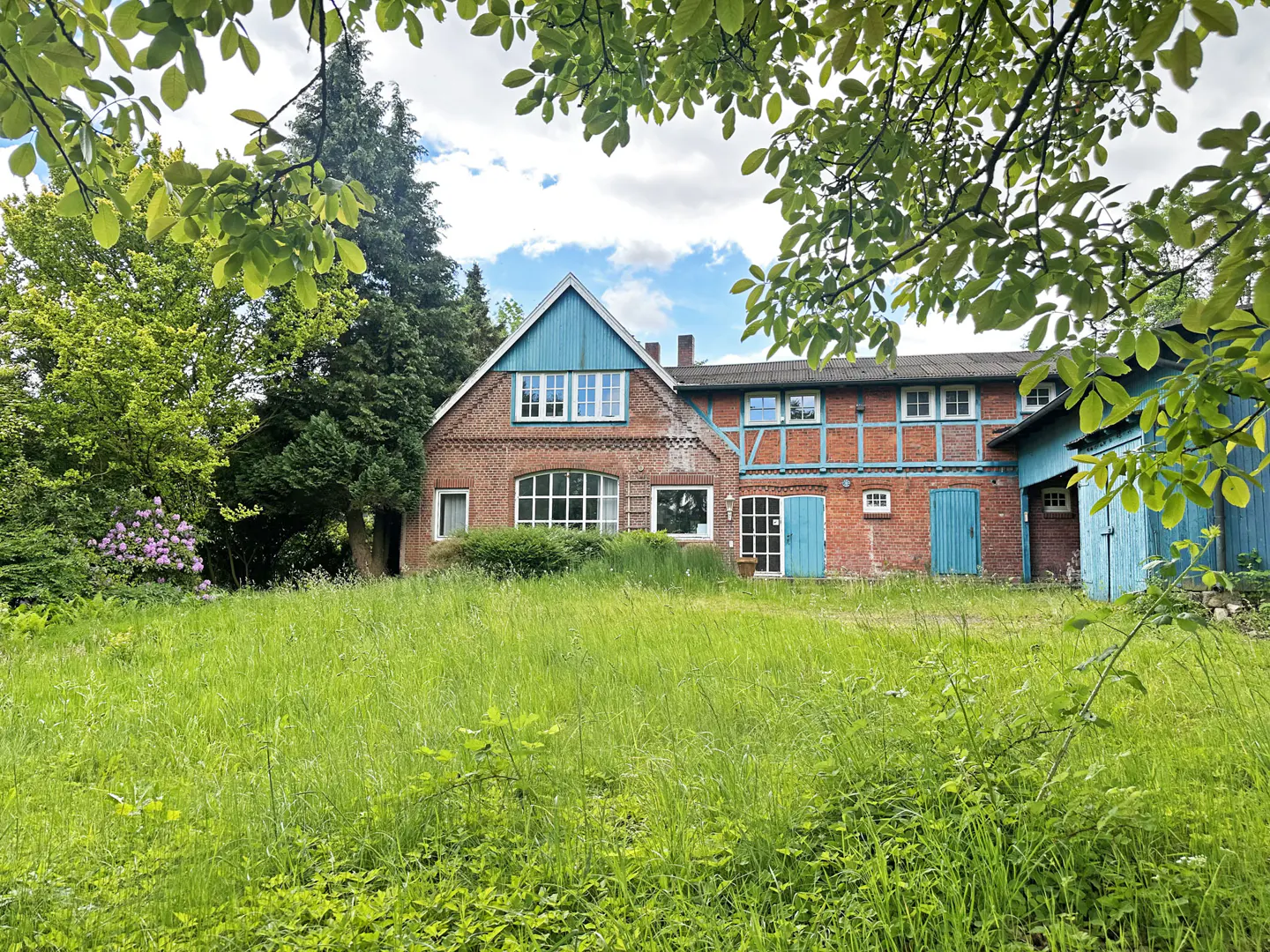 A rustic brick farmhouse with blue trim and a large grassy yard, framed by lush green trees under a cloudy sky.