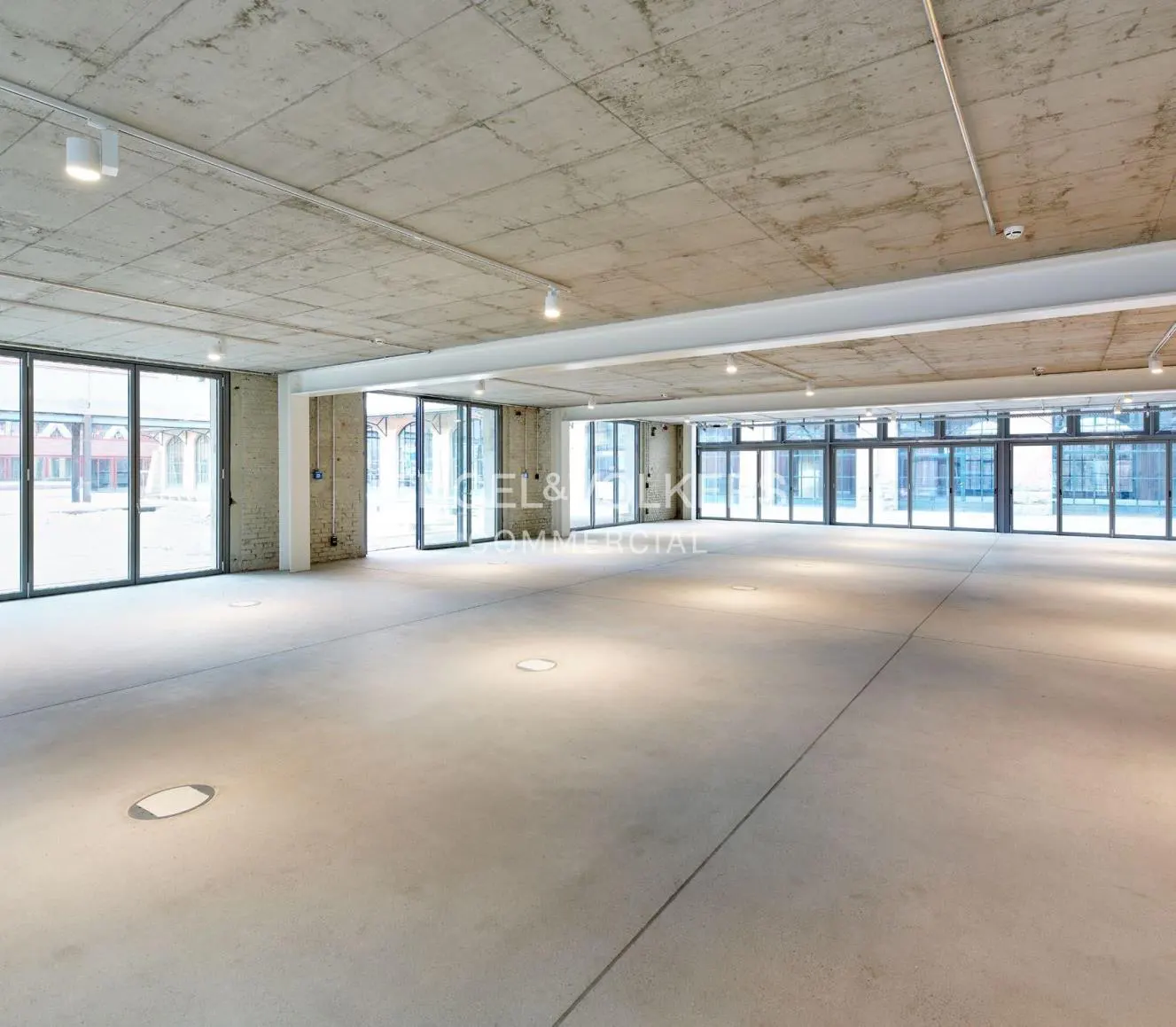 A large, empty commercial space with concrete floors and ceiling, and many windows.