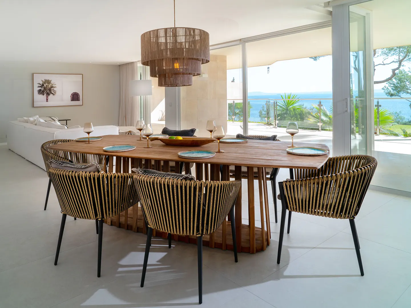 Bright dining room with a long wooden table, wicker chairs, and a tiered chandelier. Ocean view through sliding glass doors.