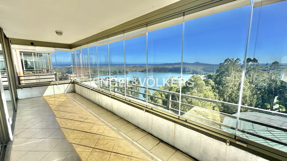A sunlit balcony with glass walls offers a scenic view of a lake, trees, and a clear blue sky.