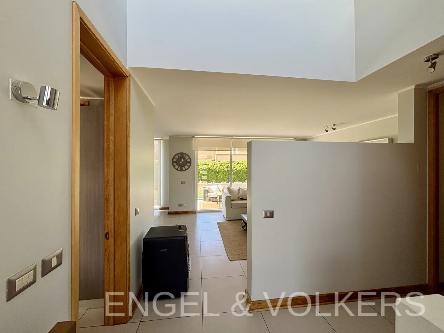 Interior view of a modern home with white walls, tile floors, and wood trim. A doorway leads to a hallway, and a living room is visible in the background.