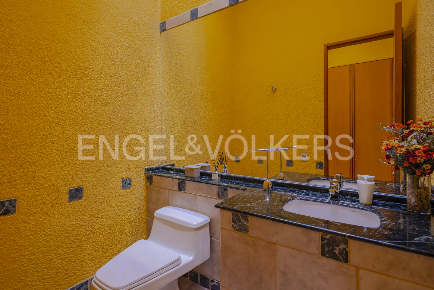 Bathroom with yellow textured walls, a marble countertop with a sink, and a white toilet. A large mirror reflects a wooden door.