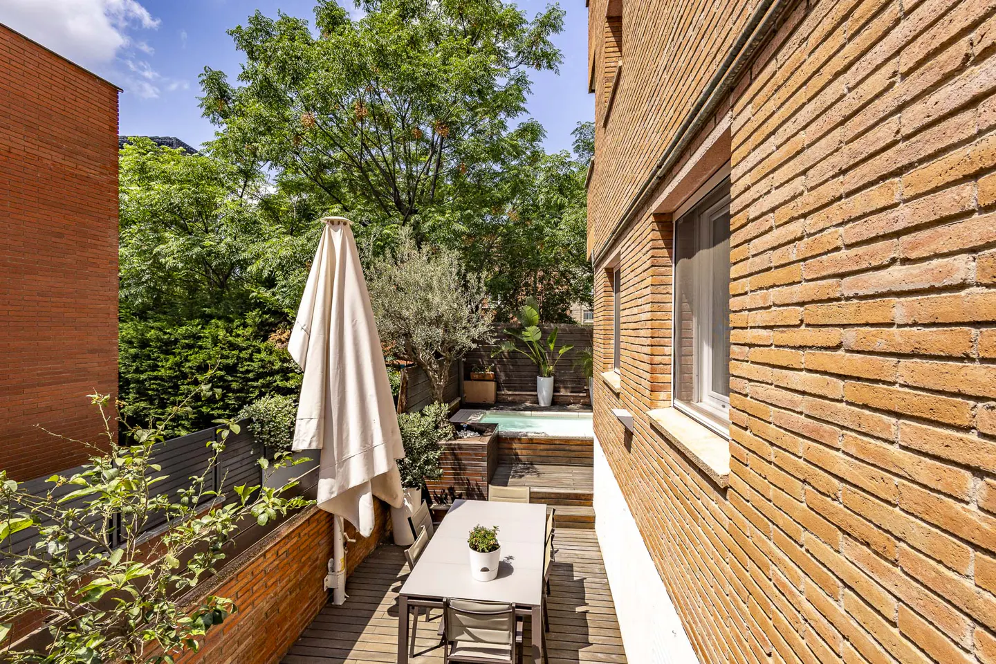 Outdoor patio with a table, chairs, and closed umbrella, next to a brick building and a small pool surrounded by greenery.