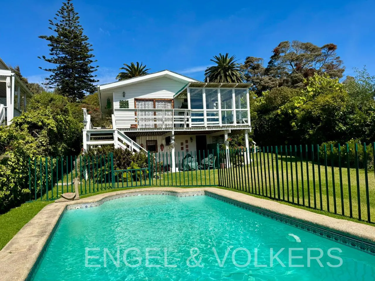 A white two-story house with a pool and green fence on a sunny day.