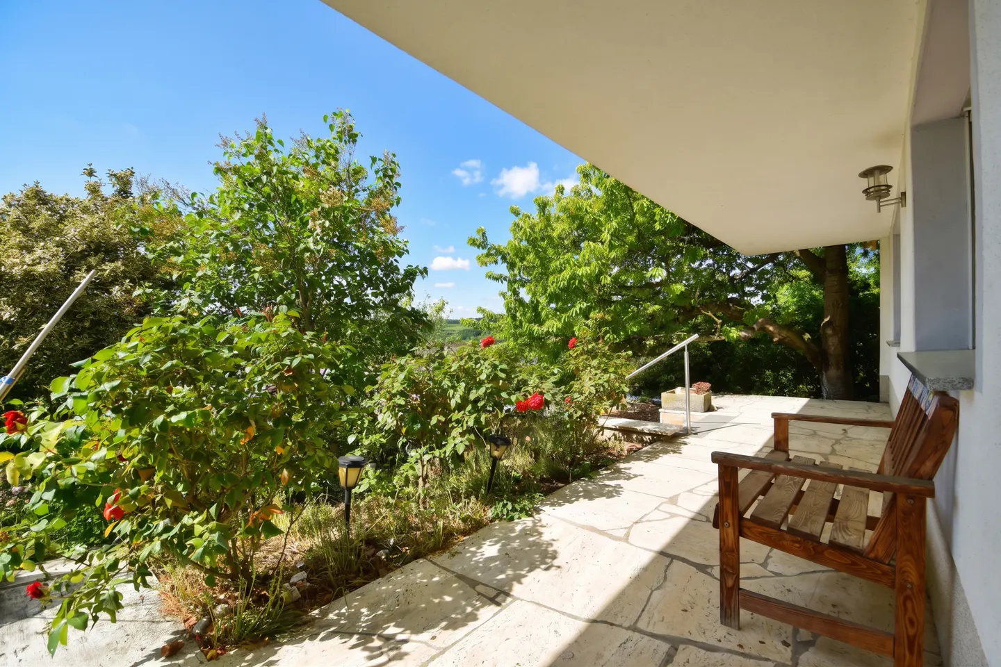 Covered patio with a wooden bench, stone floor, and lush greenery. Red roses and trees fill the background under a blue sky.