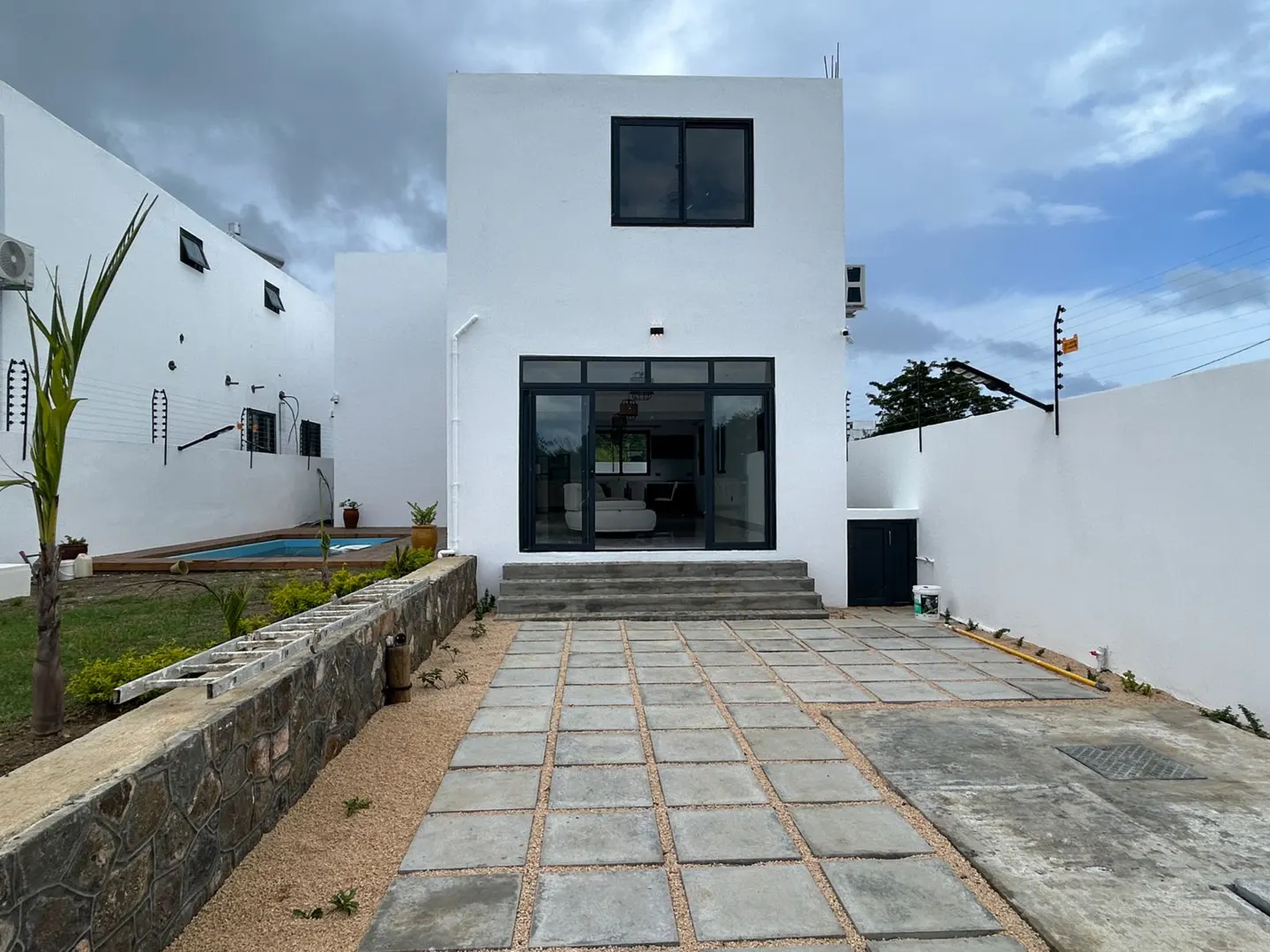 Exterior view of a modern two-story white house with black framed windows and sliding glass doors, a stone-paved patio, and a small pool in the background.