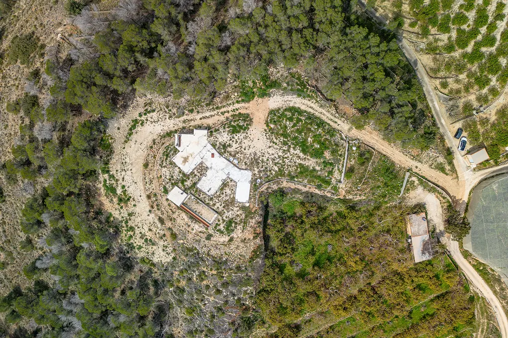 Aerial view of a white, multi-sectioned building surrounded by a dirt road and green trees.