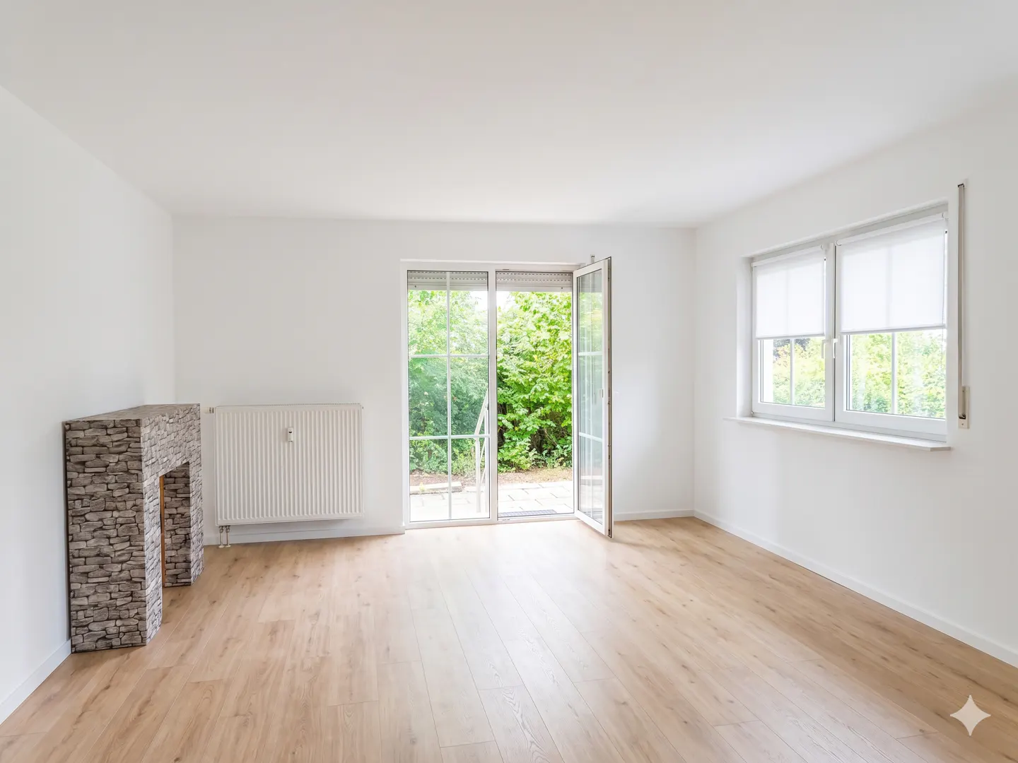 Bright, empty room with light wood floors, white walls, a stone-look fireplace, radiator, and glass doors to a garden.