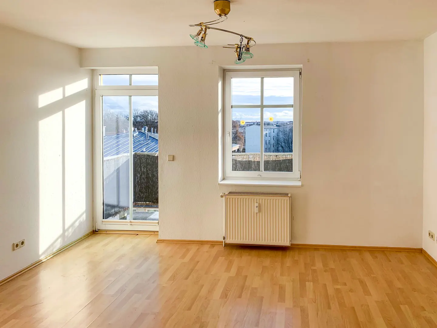 Empty room with light wood floors, white walls, a radiator, a balcony door, and a window overlooking a city view.
