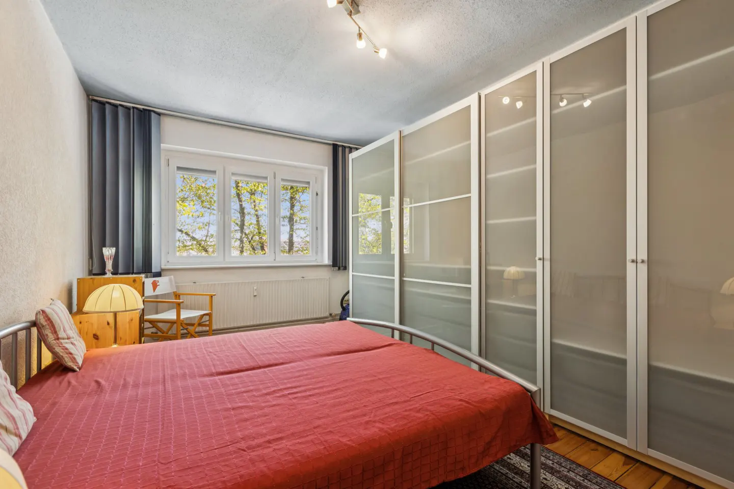 Bedroom with a red bedspread, metal frame bed, large frosted glass wardrobe, and a window with trees visible outside.