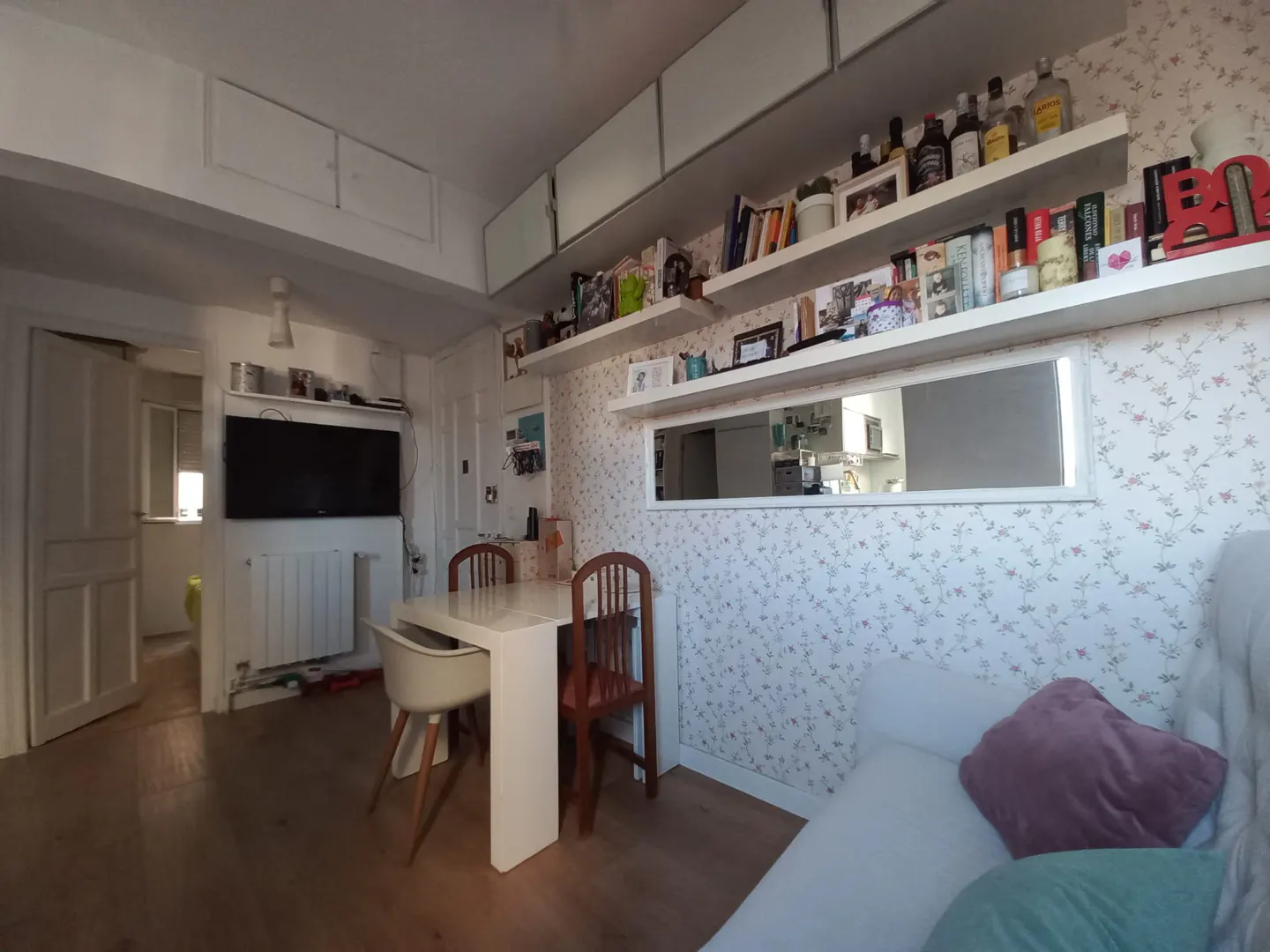 A living room with a white table and chairs, a TV, and shelves with books and bottles. The walls are covered in floral wallpaper.