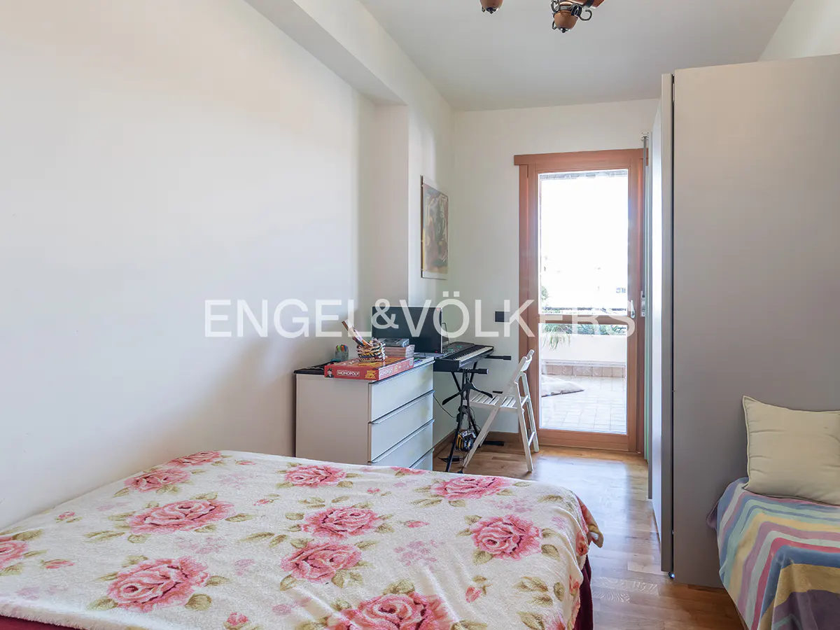 Bedroom with a bed covered in a floral pattern, a desk with a computer, and a door leading to a balcony.