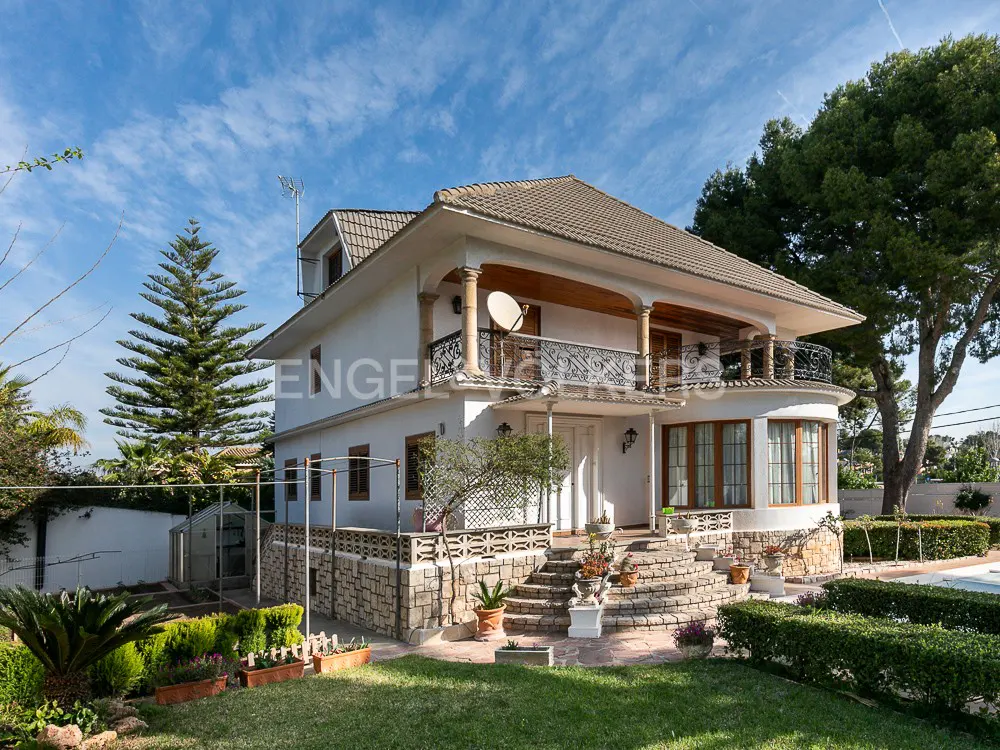 Two-story white house with a brown roof, black iron balcony, and stone base, surrounded by green lawn and trees under a blue sky.