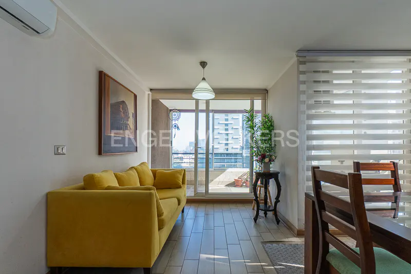 Living room with yellow sofa, wood floors, and balcony view. Dining table with chairs visible. Natural light filters through window blinds.