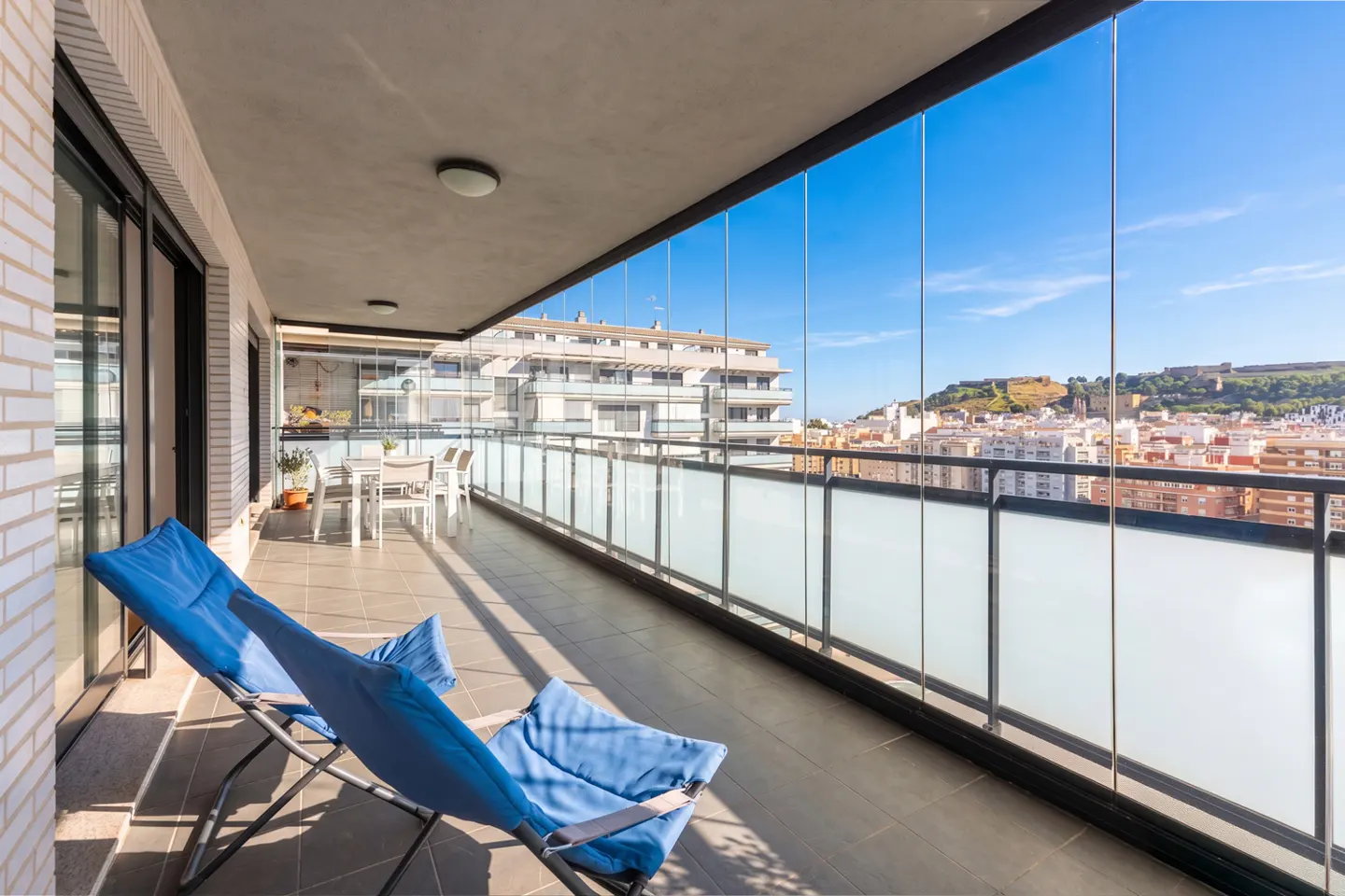 Modern balcony with blue lounge chairs, glass railings, and a panoramic view of a city and distant hillside under blue sky.