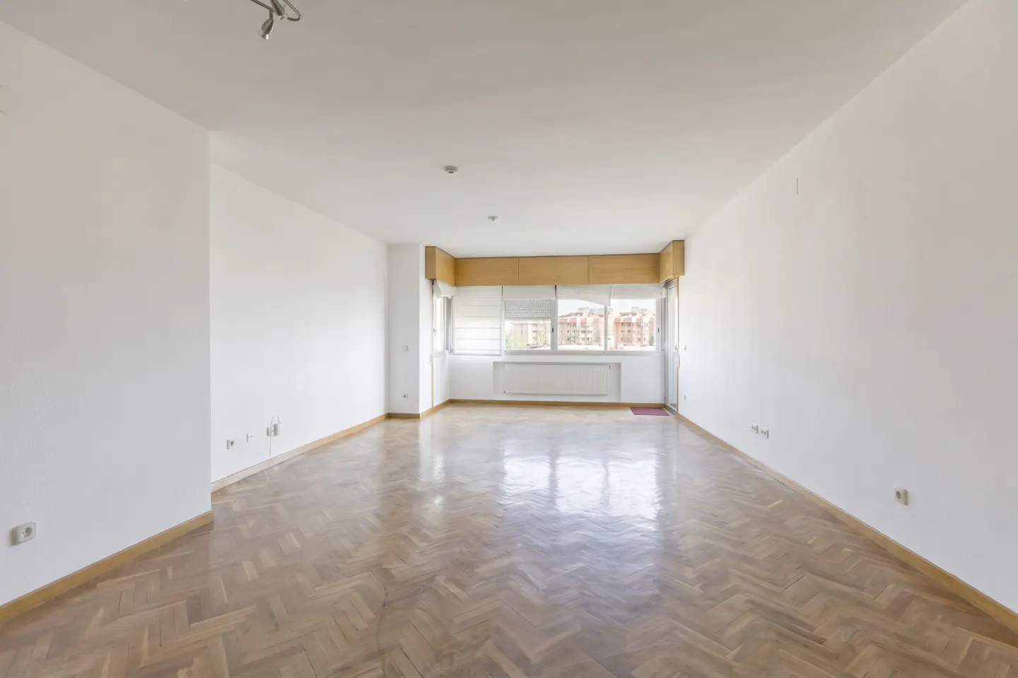 Bright, empty room with herringbone wood floors and white walls. Large window with blinds and a view of buildings.