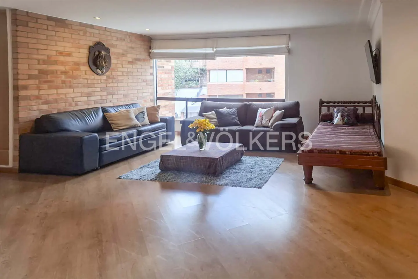 Living room with hardwood floors, brick wall, and large window. Two sofas, a wooden daybed, and a coffee table with yellow flowers.