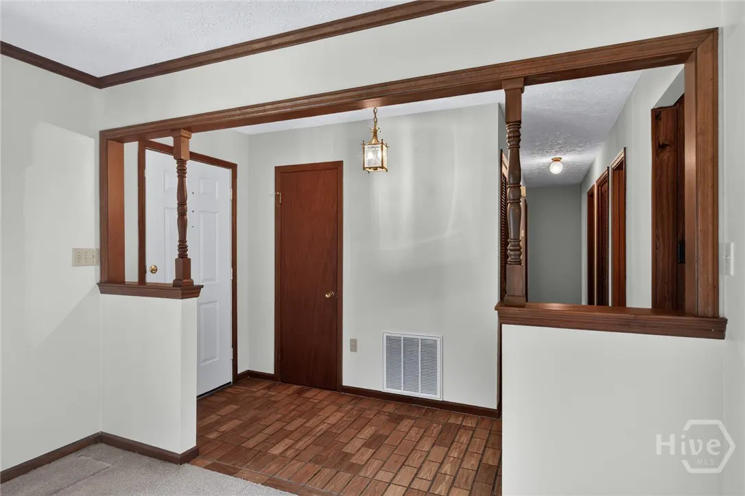 Foyer with brick floor, white walls, and dark wood trim. A white door and a brown door are visible. A hallway is seen through a wood-trimmed opening.