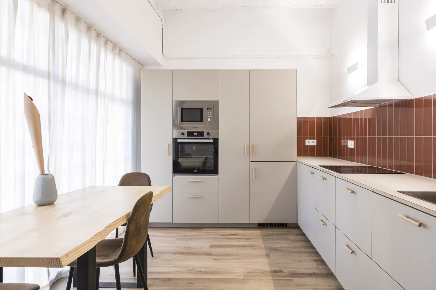 A modern kitchen with light gray cabinets, stainless steel appliances, and a wood dining table with brown chairs. A vase with a plant sits on the table.