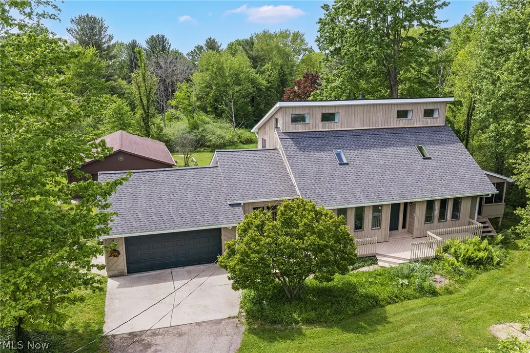 Modern two-story home with tan siding, grey shingled roof, skylights, and a dark garage, surrounded by lush green trees.