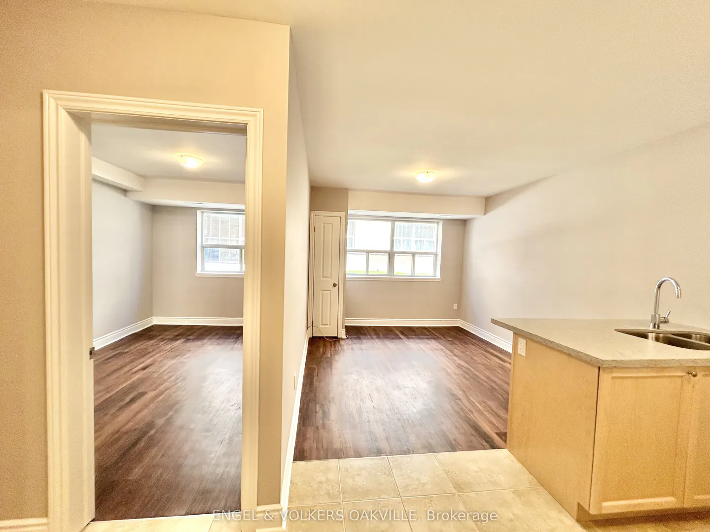A bright, empty apartment with hardwood floors, beige walls, and a kitchen island with a sink. Natural light streams through the windows.