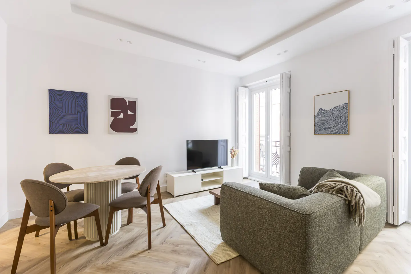 Bright living room with a round table, four chairs, a green sofa, and a TV on a white console. Art hangs on the white walls.
