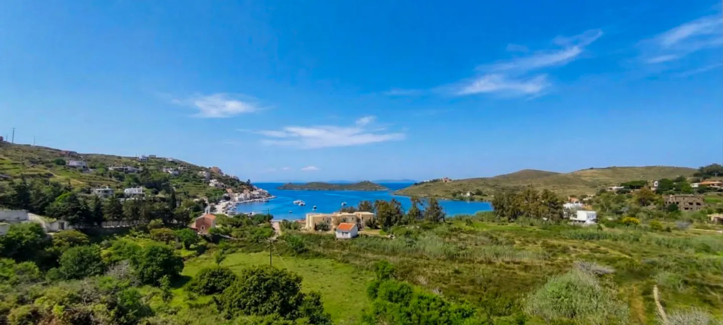 Scenic view of a Greek island with blue sea, green hills, and white houses under a bright blue sky. Boats are docked in the harbor.