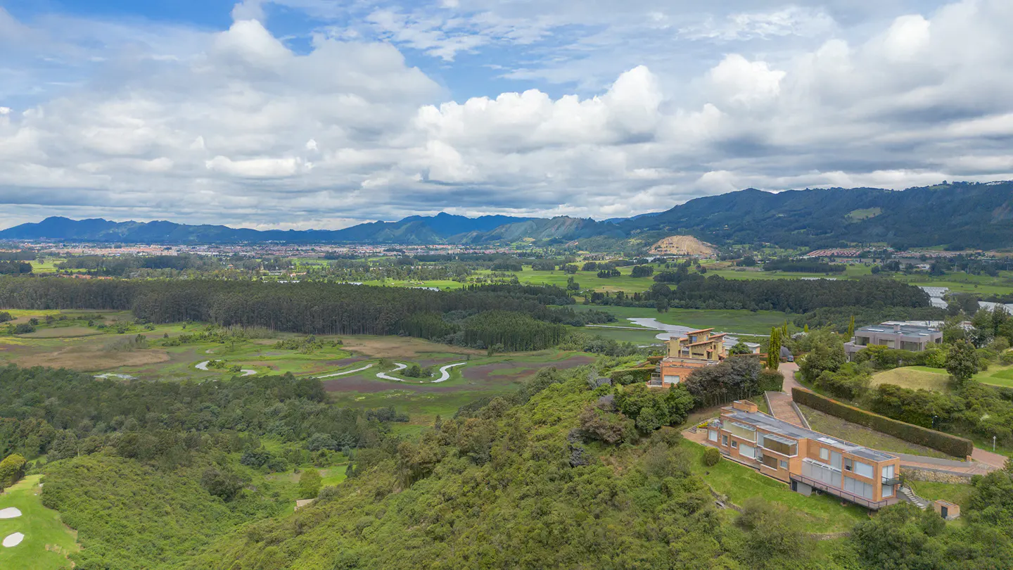 Aerial view of modern homes on a green hillside, overlooking a valley with trees, a winding river, and distant mountains under a cloudy sky.