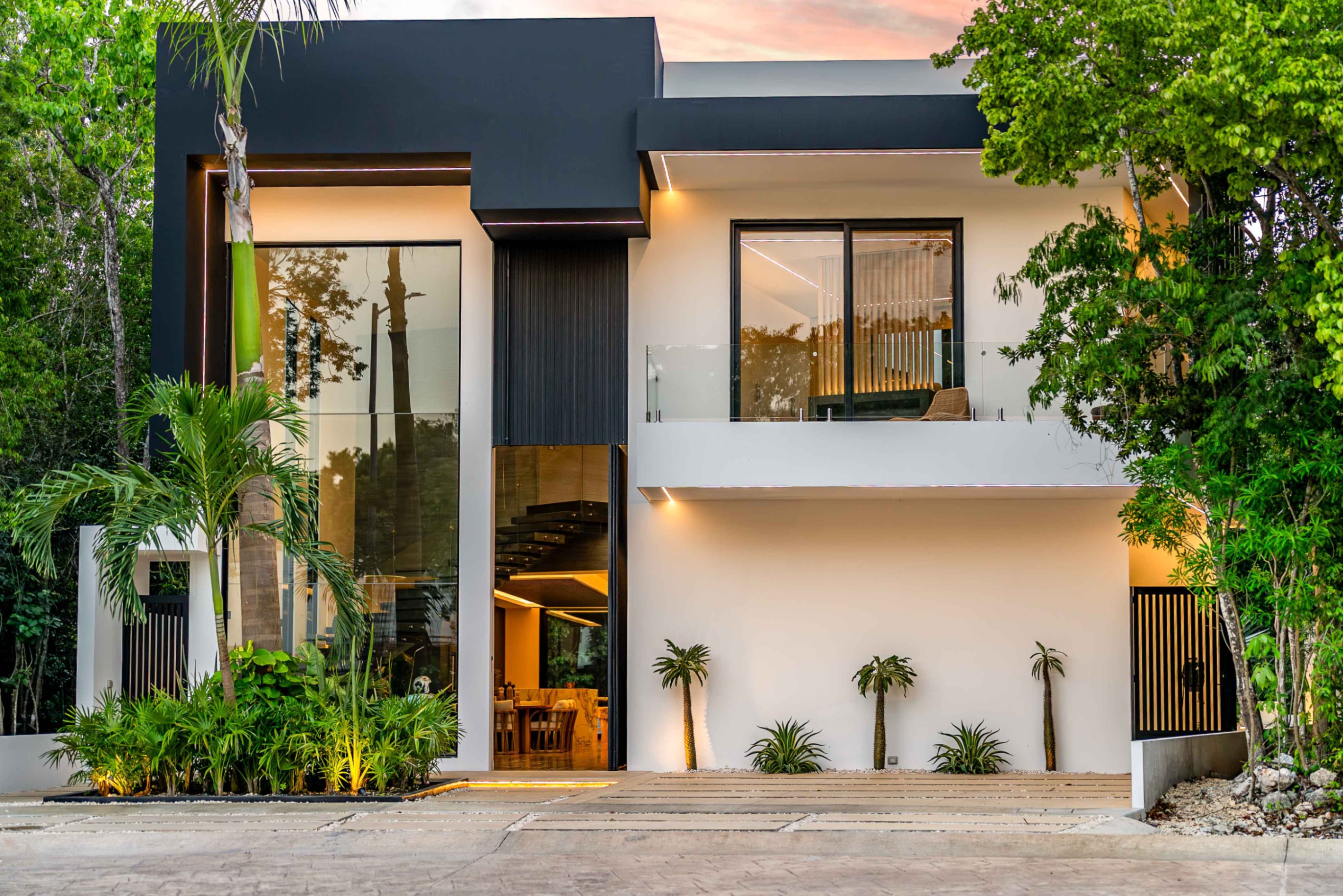 Modern two-story home with black and white exterior, large glass windows, a balcony, and lush tropical landscaping at dusk. [AI-generated text]
