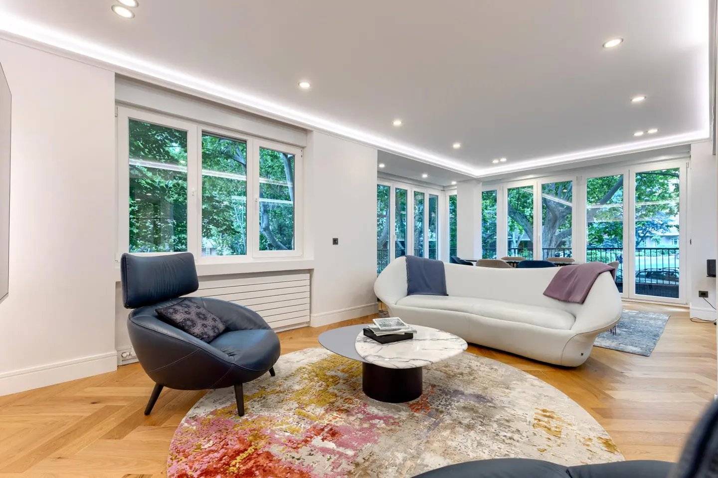 Bright living room with hardwood floors, white walls, and large windows. A blue chair and white sofa sit on a colorful rug.