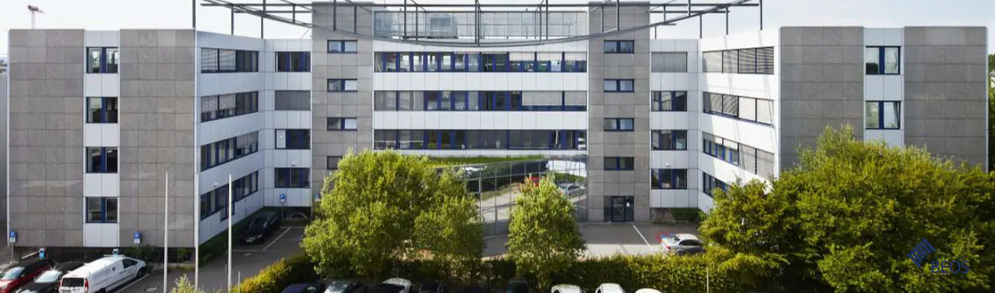 Exterior view of a modern, multi-story office building with gray and white facade, blue-framed windows, and green trees in front.