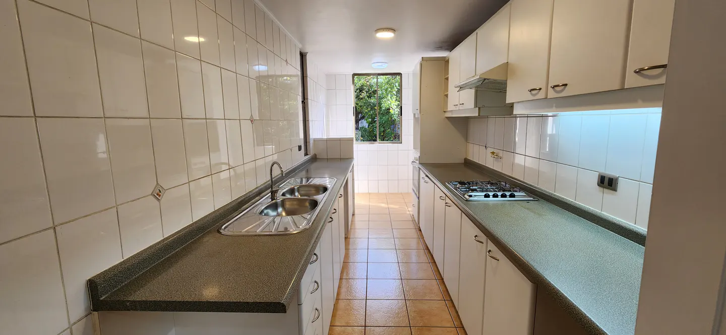 A galley kitchen with white cabinets, a stainless steel sink, and a gas stove. A window at the end of the kitchen lets in natural light.