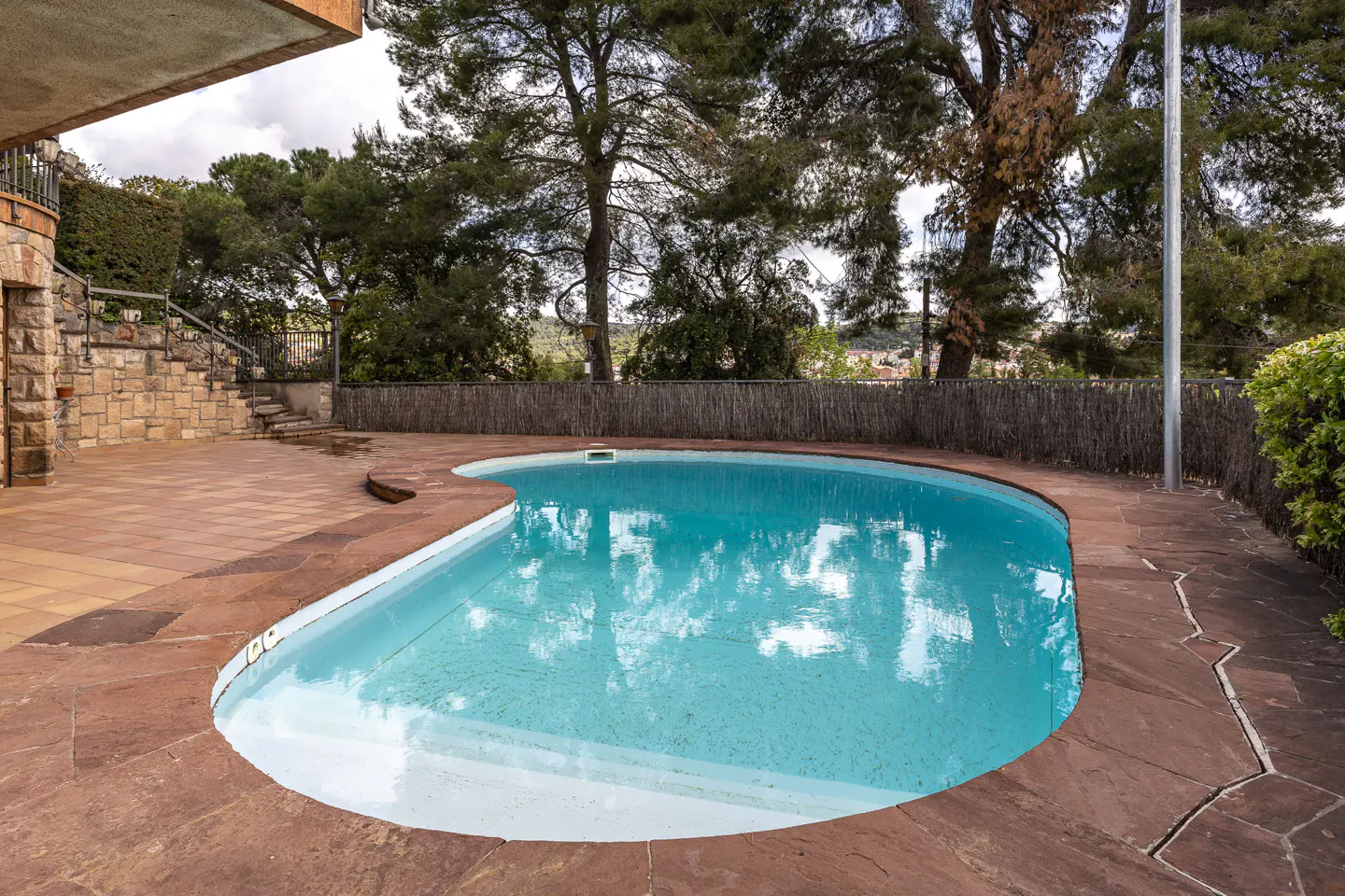 A kidney-shaped pool with blue water is surrounded by a brown stone patio and a fence with trees in the background.