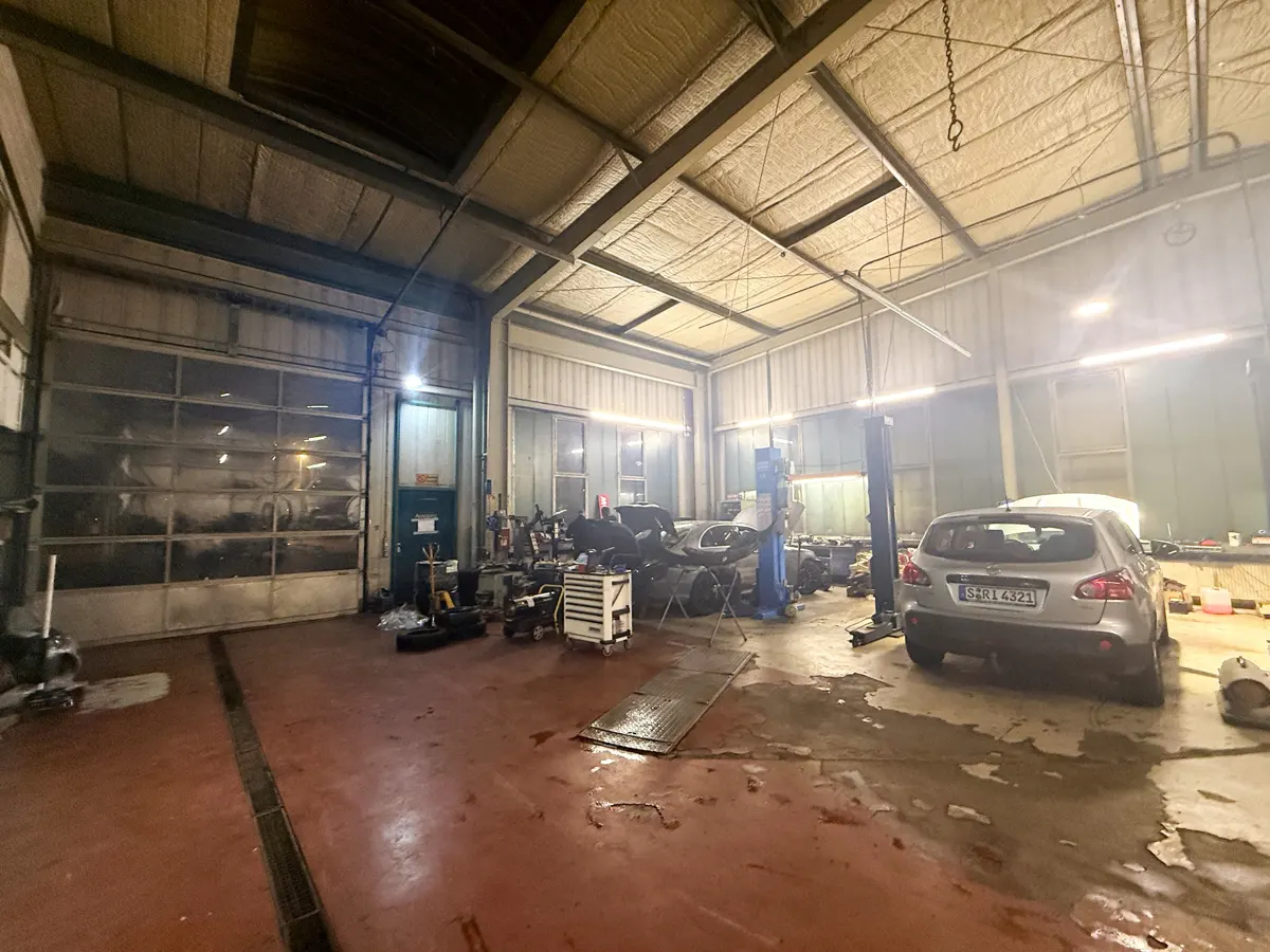 A wide shot of a large, cluttered auto repair shop with a gray car parked inside. The shop has a high ceiling, a large garage door, and various tools and equipment.