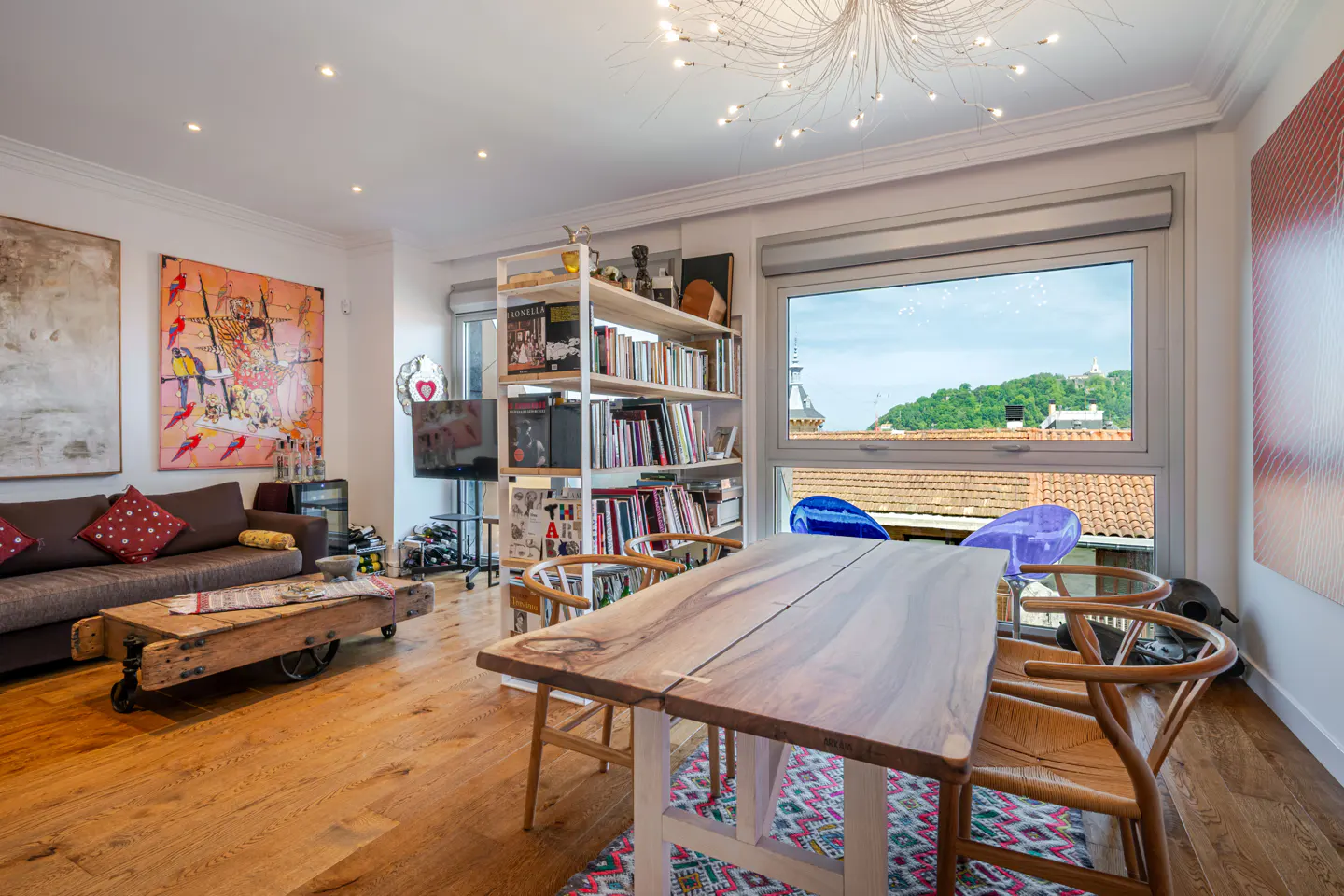 Bright living room with wood floors, a brown sofa, and a rustic wood table. A large window overlooks a cityscape. Colorful art adorns the walls.
