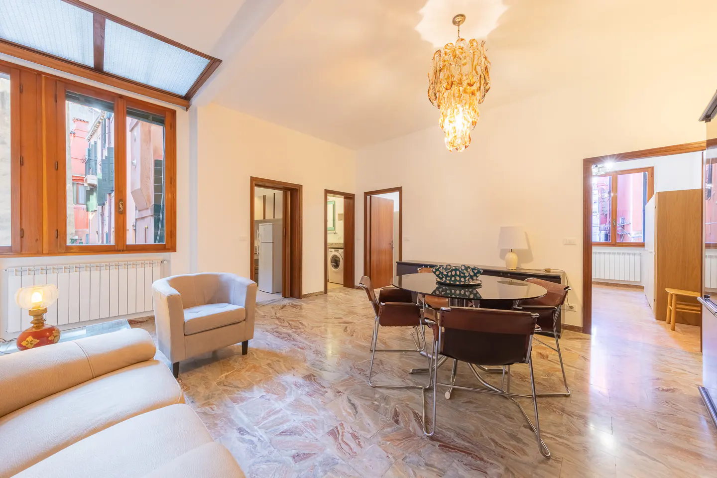 Bright living room with marble floors, beige sofa and armchair, dining table with chairs, and a view of a Venice canal through a window.
