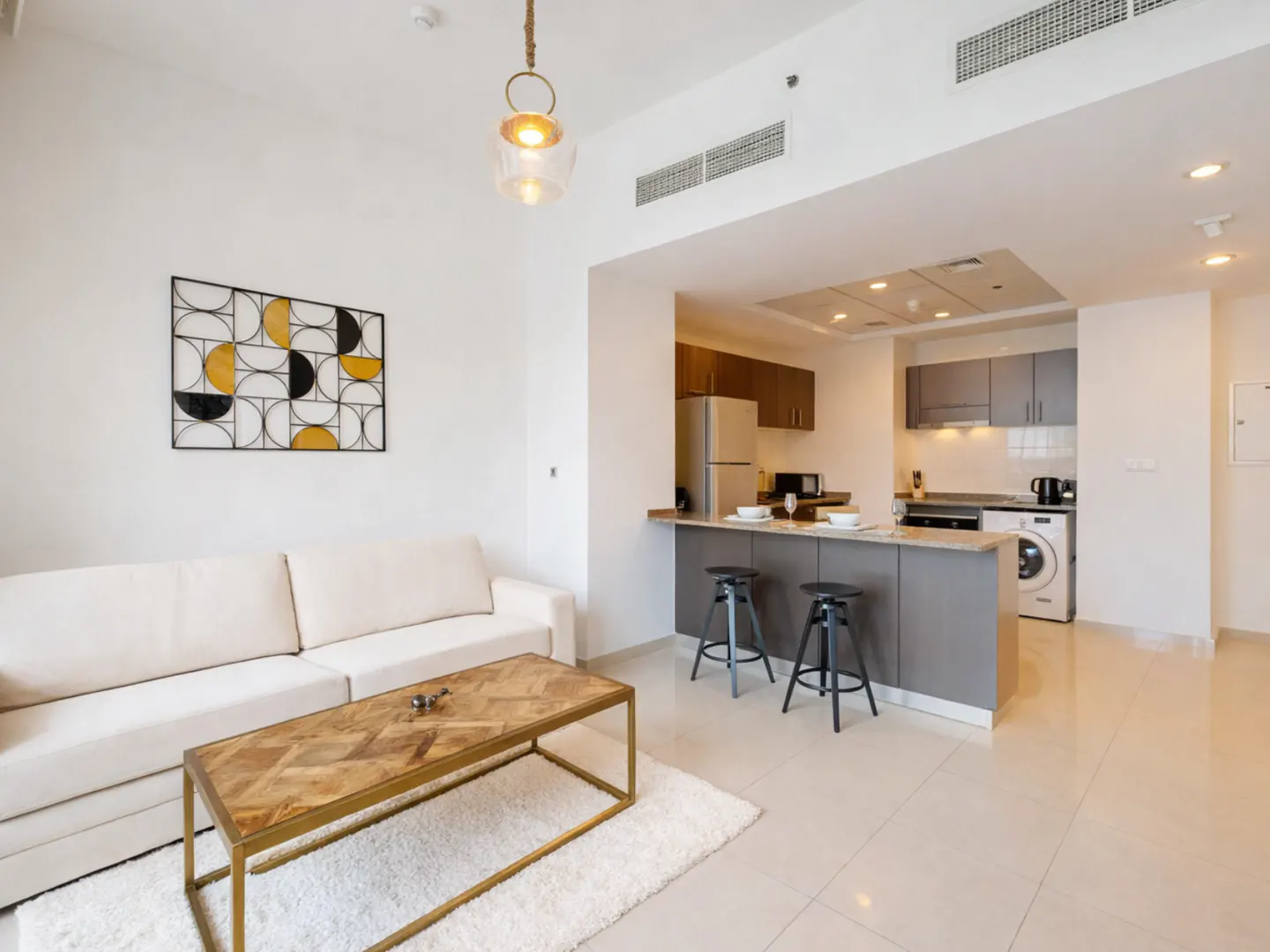 A bright, modern apartment interior with a white sofa, wood table, and open kitchen with bar stools.