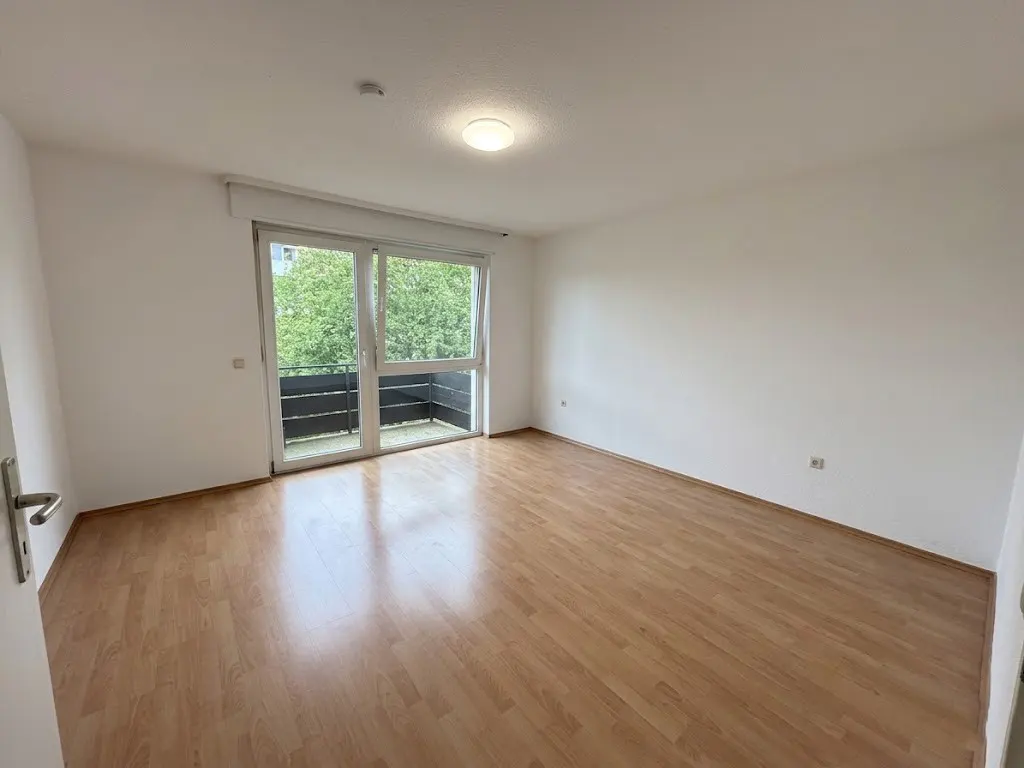 Empty room with light wood floor, white walls, and glass doors to a balcony with green trees visible outside.