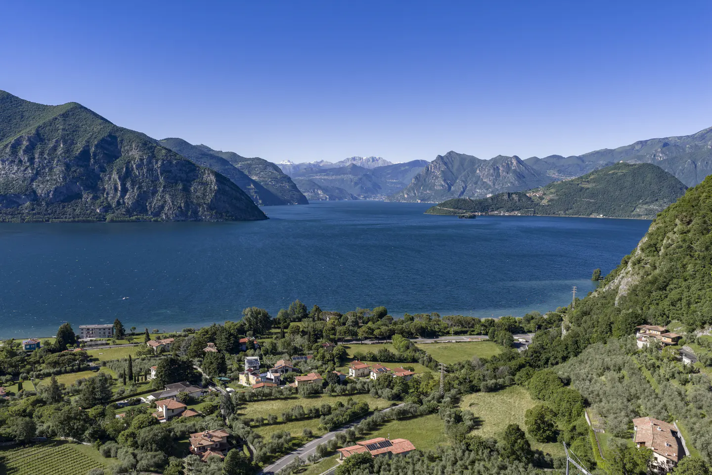 Scenic view of Lake Iseo, Italy, with blue water, green hills, and red-roofed houses under a clear blue sky.