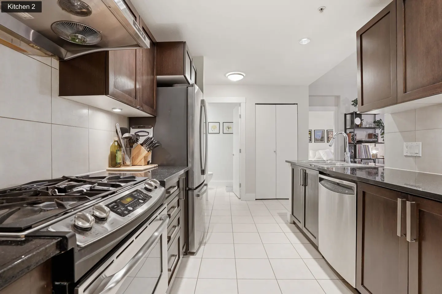 A modern kitchen with stainless steel appliances, dark wood cabinets, and white tile flooring. A gas range stove is visible on the left.