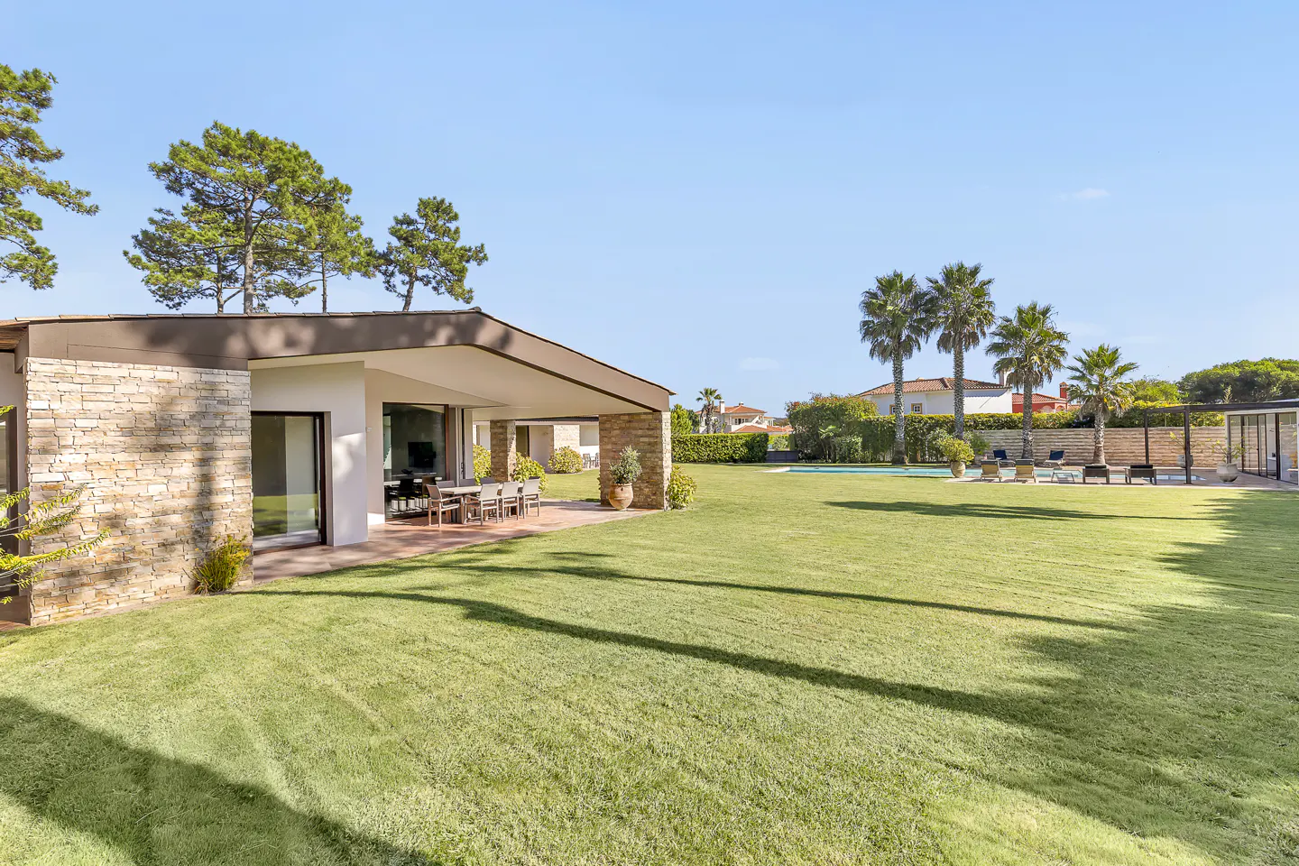 Backyard view of a modern house with a stone wall, patio with table, green lawn, pool, palm trees, and blue sky.